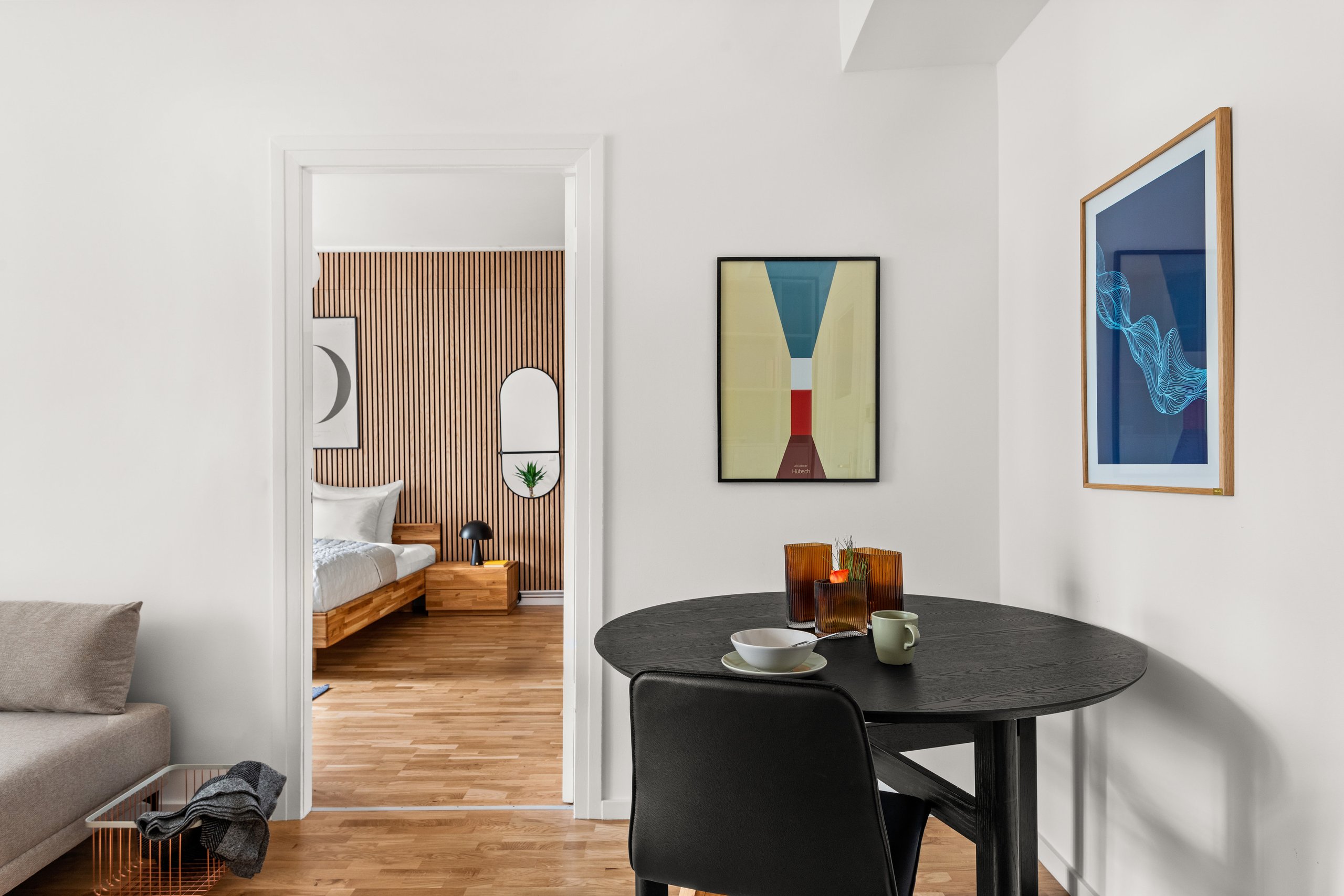 Blick durch eine offene Tür von einem Essbereich mit schwarzem Tisch in ein Schlafzimmer mit Holzbett und vertikaler Holzverkleidung.