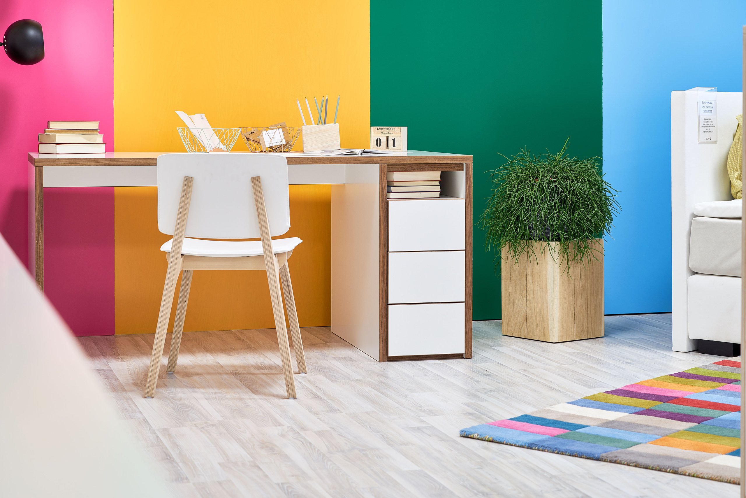 Holzconnection: Ein Schreibtisch mit Stuhl, Pflanzen und Teppich vor einer Wand in Pink, Gelb, Grün und Blau