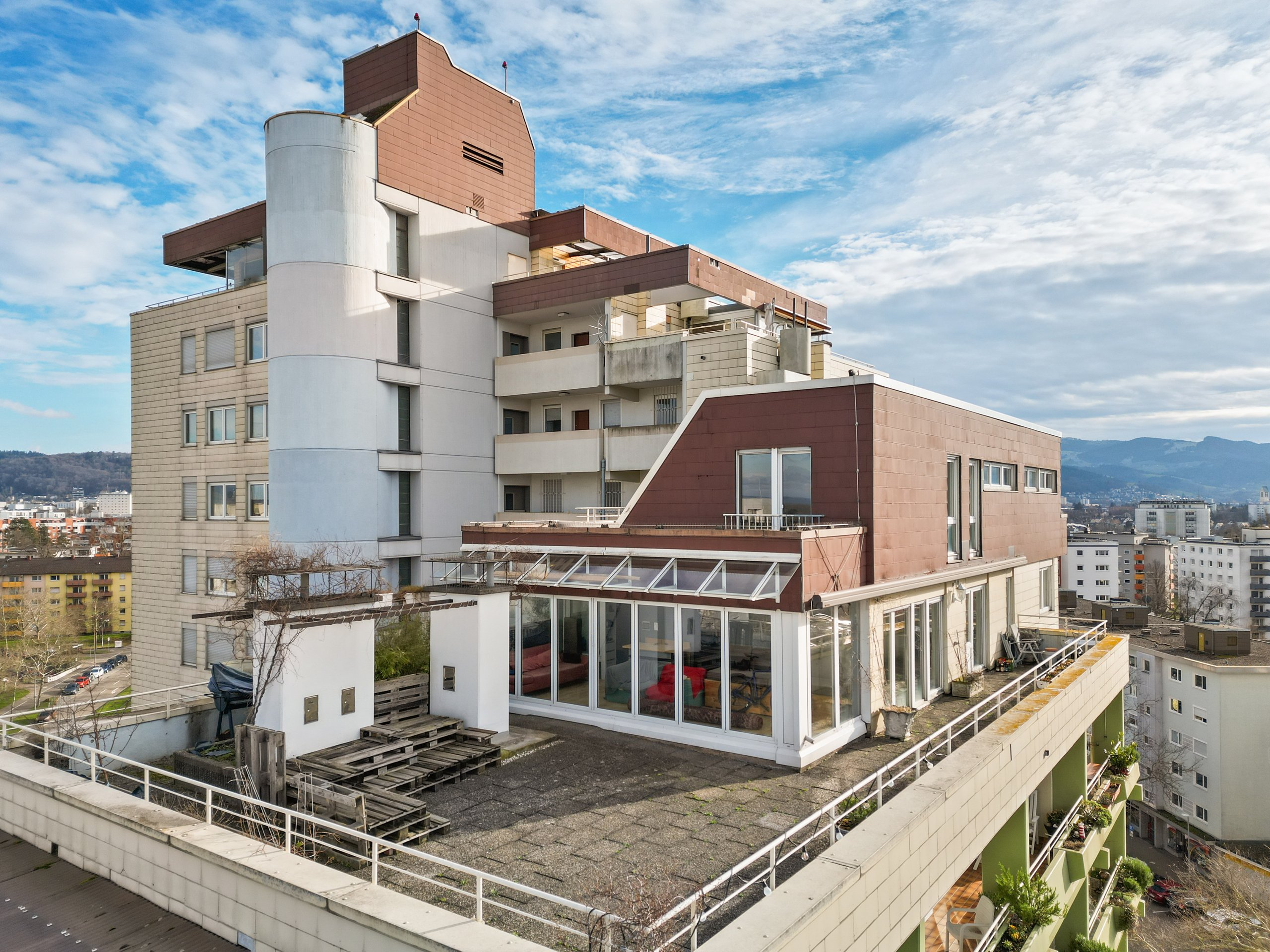Mehrstöckiges Wohngebäude mit braunen und weißen Fassaden, Balkonen und einer Dachterrasse mit Wintergarten unter blauem Himmel.