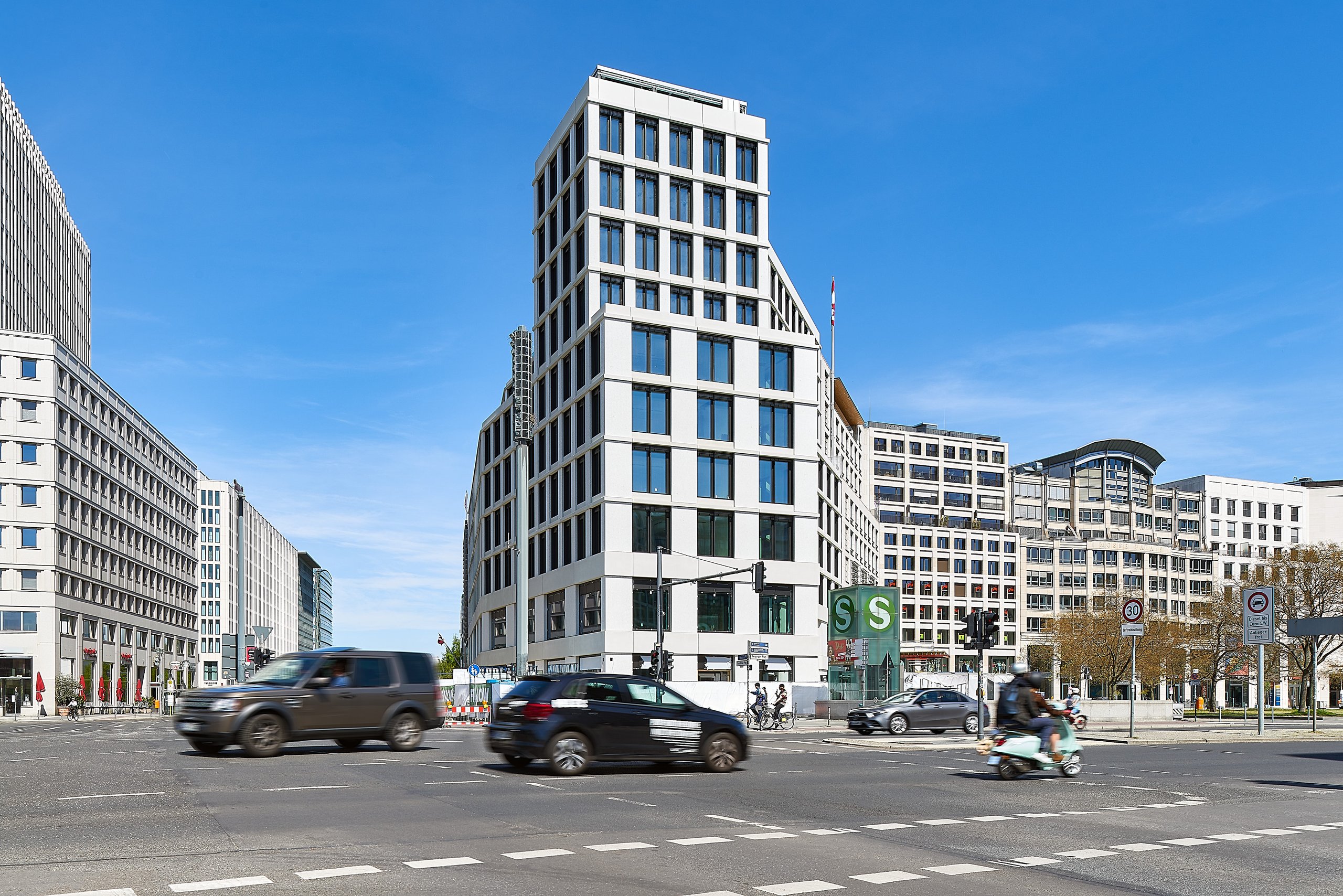 TRION Leipziger Platz Berlin, für AGIB Real Estate: Mehrere Autos und ein Roller fahren auf einer breiten Straße vor modernen Gebäuden unter blauem Himmel