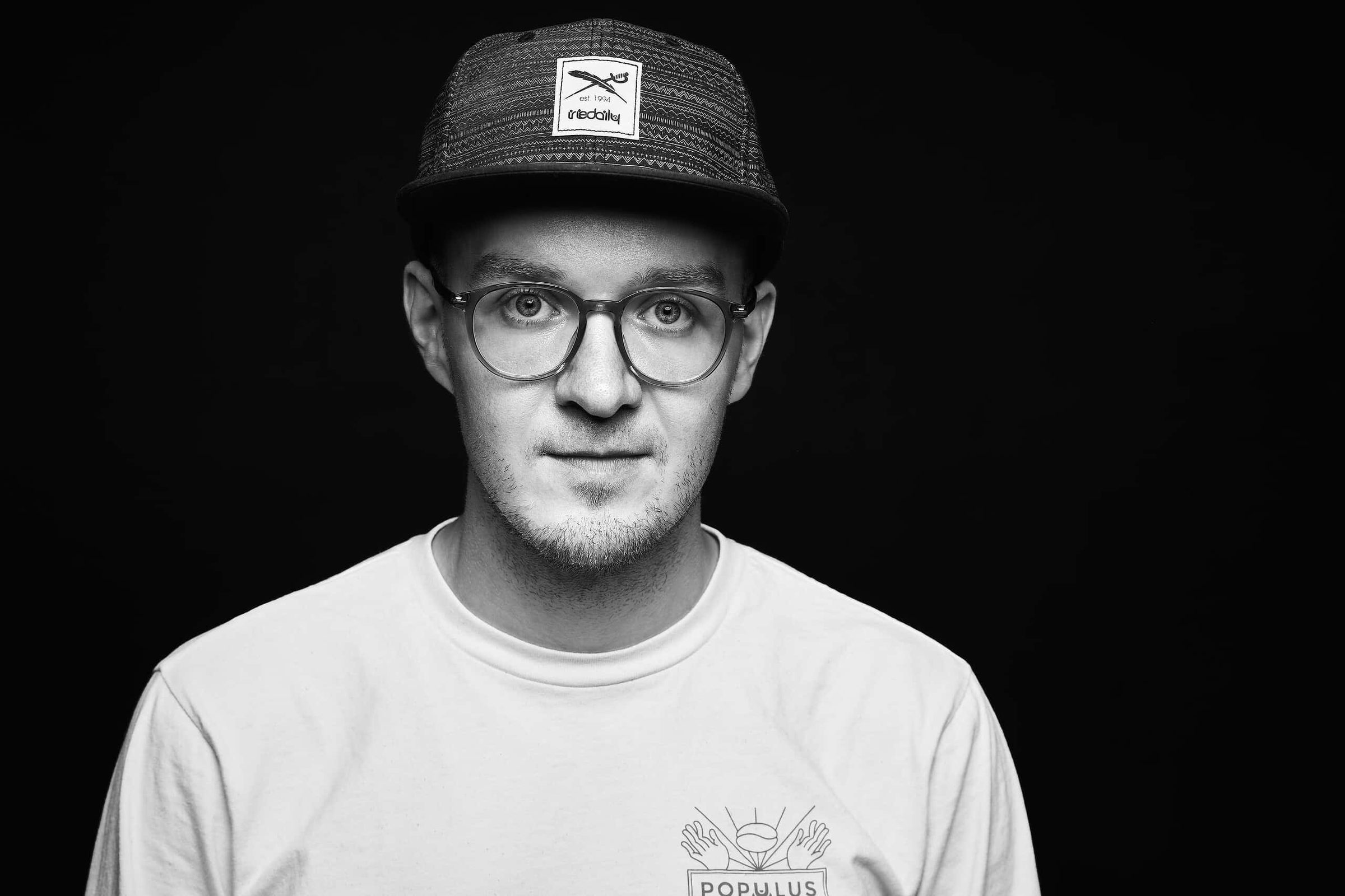 Thomas Stibenz, Businessfotograf und Architekturfotograf aus Freiburg, Schwarz-Weiß-Porträt mit Brille, Bart und Kappe vor schwarzem Hintergrund
